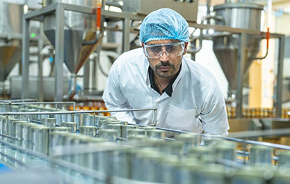 Eine Person in Schutzkleidung, Haube und Schutzbrille beobachtet eine Produktionslinie mit Metallbehältern oder Lebensmitteldosen. Im Hintergrund sind Maschinen und industrielle Anlagen zu sehen.