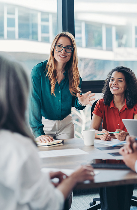 Mehrere Personen sitzen an einem Tisch in einem Besprechungsraum. Eine Frau steht und spricht, w&auml;hrend die anderen zuh&ouml;ren, Notizen machen oder auf einen Laptop schauen. Die Atmosph&auml;re ist kooperativ.