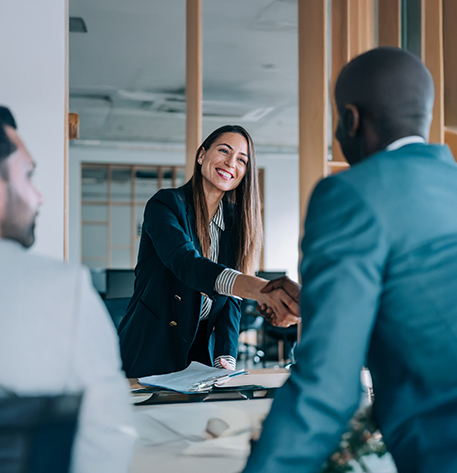 Eine Frau steht an einem Tisch in einem B&uuml;ro und reicht einer anderen Person die Hand. Zwei weitere Personen sitzen am Tisch und beobachten die Begr&uuml;&szlig;ung. Die Stimmung wirkt offen und professionell.