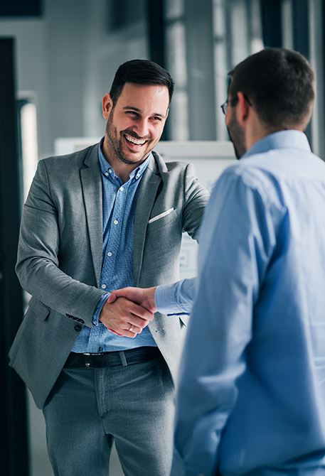 Zwei M&auml;nner stehen in einem B&uuml;roflur und geben sich die Hand. Beide l&auml;cheln, die Situation wirkt freundlich und professionell.