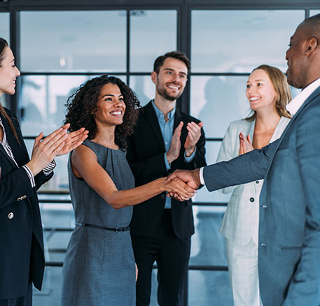 Mehrere Personen stehen in einem B&uuml;ro zusammen. Zwei Personen geben sich die Hand, w&auml;hrend die anderen klatschen und l&auml;cheln. Die Szene zeigt Anerkennung und Teamgeist.