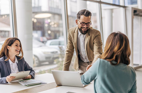 Drei Personen sitzen an einem hellen Bürotisch. Eine Person steht leicht nach vorne gebeugt und lächelt, während sie einer sitzenden Person die Hand schüttelt. Die Gruppe führt ein freundliches, geschäftliches Gespräch, Laptops und Unterlagen liegen auf dem Tisch.