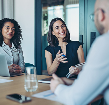 Drei Personen sitzen an einem Tisch in einem Besprechungsraum. Eine Frau spricht und gestikuliert, die anderen h&ouml;ren aufmerksam zu. Auf dem Tisch liegen Unterlagen und ein Laptop.