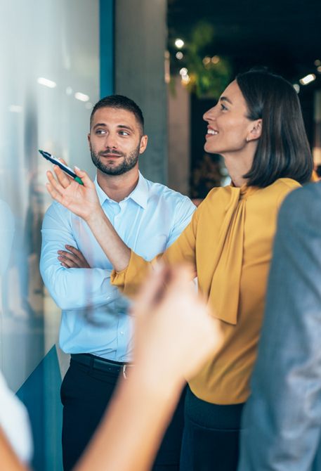 Mehrere Personen stehen vor einer Glas- oder Whiteboardfl&auml;che. Eine Frau zeigt mit einem Stift auf Inhalte, ein Mann h&ouml;rt aufmerksam zu. Die Gruppe diskutiert gemeinsam.