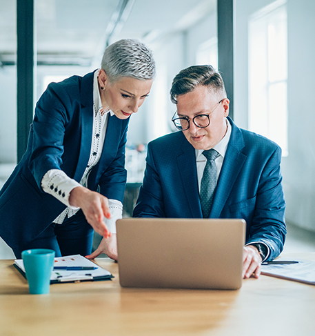 Eine Frau und ein Mann in Businesskleidung sitzen an einem Tisch und schauen auf einen Laptop. Die Frau deutet auf den Bildschirm, beide wirken konzentriert.
