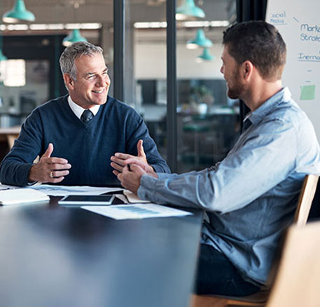Zwei Personen sitzen einander an einem langen Tisch gegenüber und führen ein konzentriertes, freundliches Gespräch. Unterlagen und ein Tablet liegen auf dem Tisch, im Hintergrund ist ein modern gestalteter Bürobereich zu sehen.