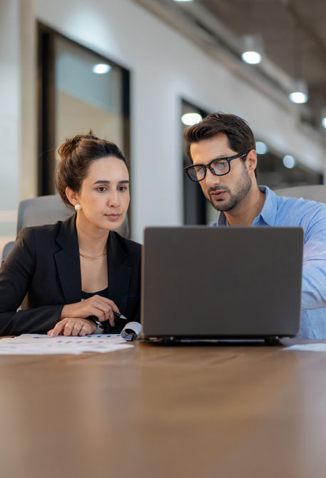 Zwei Personen sitzen nebeneinander an einem Tisch und schauen gemeinsam auf einen Laptop. Vor ihnen liegen verschiedene Unterlagen. Die Umgebung wirkt wie ein modernes Großraumbüro.