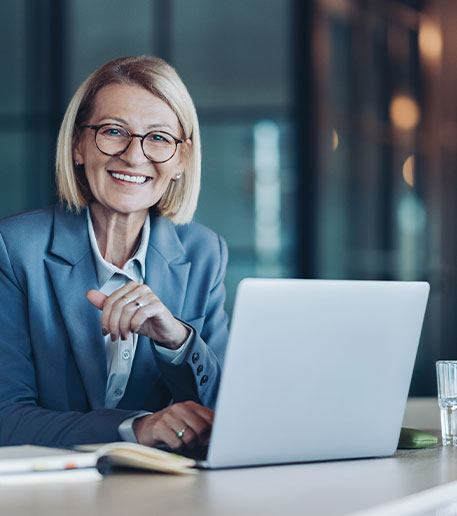 Eine l&auml;chelnde Gesch&auml;ftsfrau sitzt mit Laptop am Schreibtisch und vermittelt Kompetenz, Erfahrung und eine positive Arbeitsatmosph&auml;re.