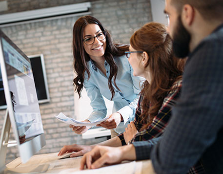 Eine Frau steht an einem Schreibtisch und zeigt zwei sitzenden Personen Unterlagen auf einem gro&szlig;en Monitor. Alle drei tauschen sich &uuml;ber die angezeigten Inhalte aus und arbeiten gemeinsam an einer Aufgabe.