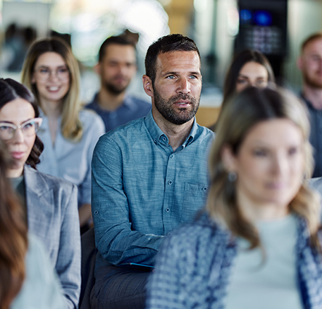 Mehrere Personen sitzen in Reihen und h&ouml;ren aufmerksam zu. Im Vordergrund ist ein Mann zu sehen, der konzentriert nach vorne blickt, w&auml;hrend weitere Teilnehmende unscharf im Hintergrund sitzen.