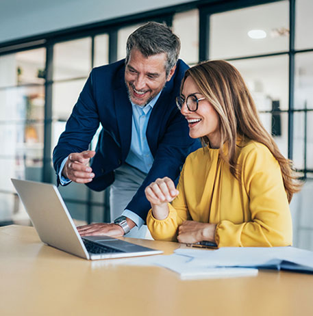 Ein Mann und eine Frau sitzen an einem Tisch und arbeiten gemeinsam an einem Laptop. Der Mann zeigt auf den Bildschirm, die Frau l&auml;chelt und h&ouml;rt aufmerksam zu. Die Situation wirkt unterst&uuml;tzend und kooperativ.